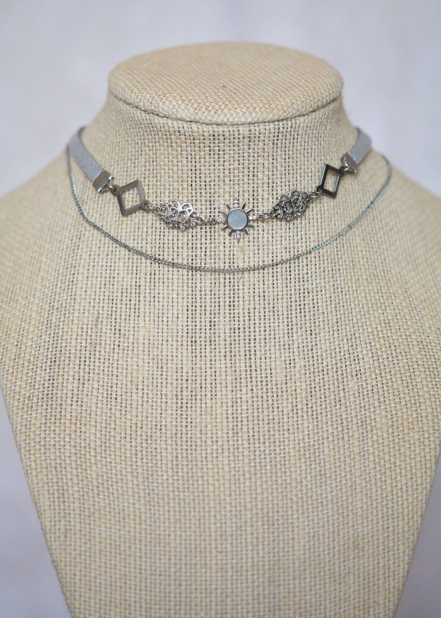 Collier en suède argenté avec soleil émaillé blanc, strass et détails en acier. Bijou léger d’inspiration céleste et angélique.