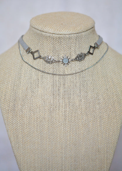 Collier en suède argenté avec soleil émaillé blanc, strass et détails en acier. Bijou léger d’inspiration céleste et angélique.