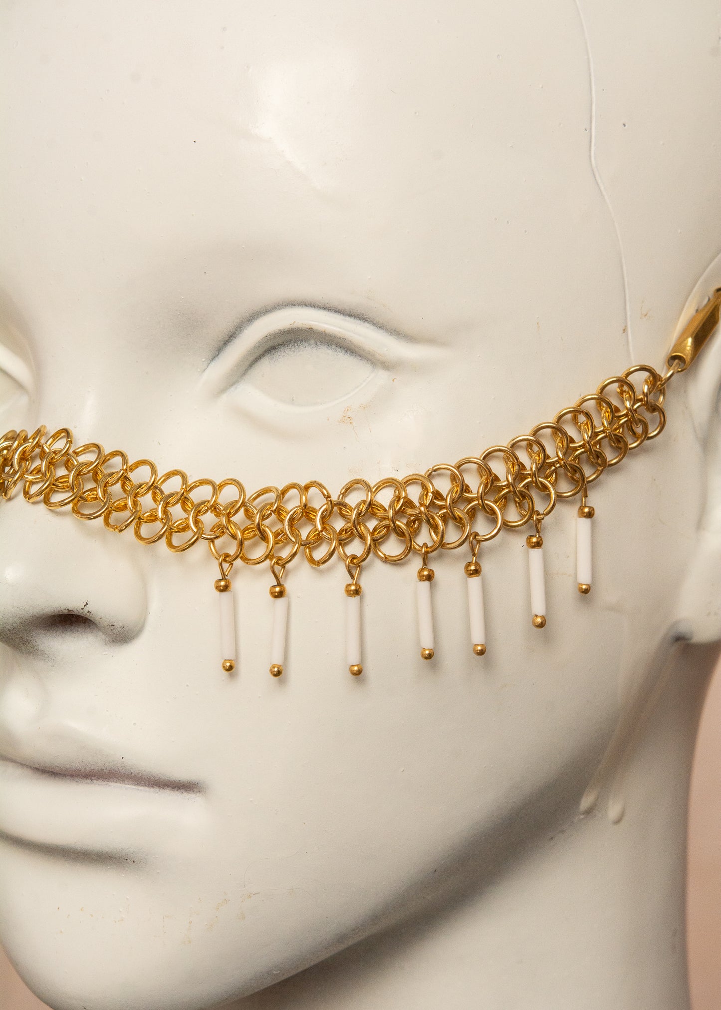 Chaîne de visage en acier inoxydable, assemblée à la main dans un motif de côte-de-maille avec perles tubes blanches et perles de lave dorées. Bijou mystique et guerrier, parfait pour les festivals et événements, ou pour ajouter une touche orientale à un look audacieux.