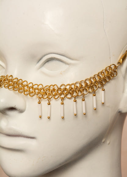 Chaîne de visage en acier inoxydable, assemblée à la main dans un motif de côte-de-maille avec perles tubes blanches et perles de lave dorées. Bijou mystique et guerrier, parfait pour les festivals et événements, ou pour ajouter une touche orientale à un look audacieux.
