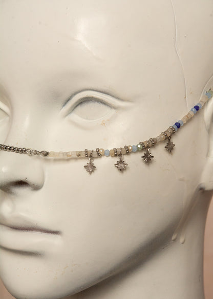 Bijou facial artisanal combinant agate blanche, cristaux bleus et acier inoxydable. Accessoire céleste avec charmes étoiles et pierres de lune noires aux reflets holographiques transformées en lanternes orientales. Pièce unique apportant sérénité et équilibre, parfaite pour festivals nocturnes et événements mystiques.