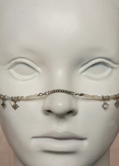Bijou facial artisanal combinant agate blanche, cristaux bleus et acier inoxydable. Accessoire céleste avec charmes étoiles et pierres de lune noires aux reflets holographiques transformées en lanternes orientales. Pièce unique apportant sérénité et équilibre, parfaite pour festivals nocturnes et événements mystiques.